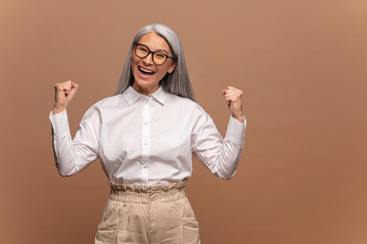 Portrait of excited overjoyed business woman with grey hair standing with raised fists and shouting yeah, I'm a winner, rejoicing victory, success